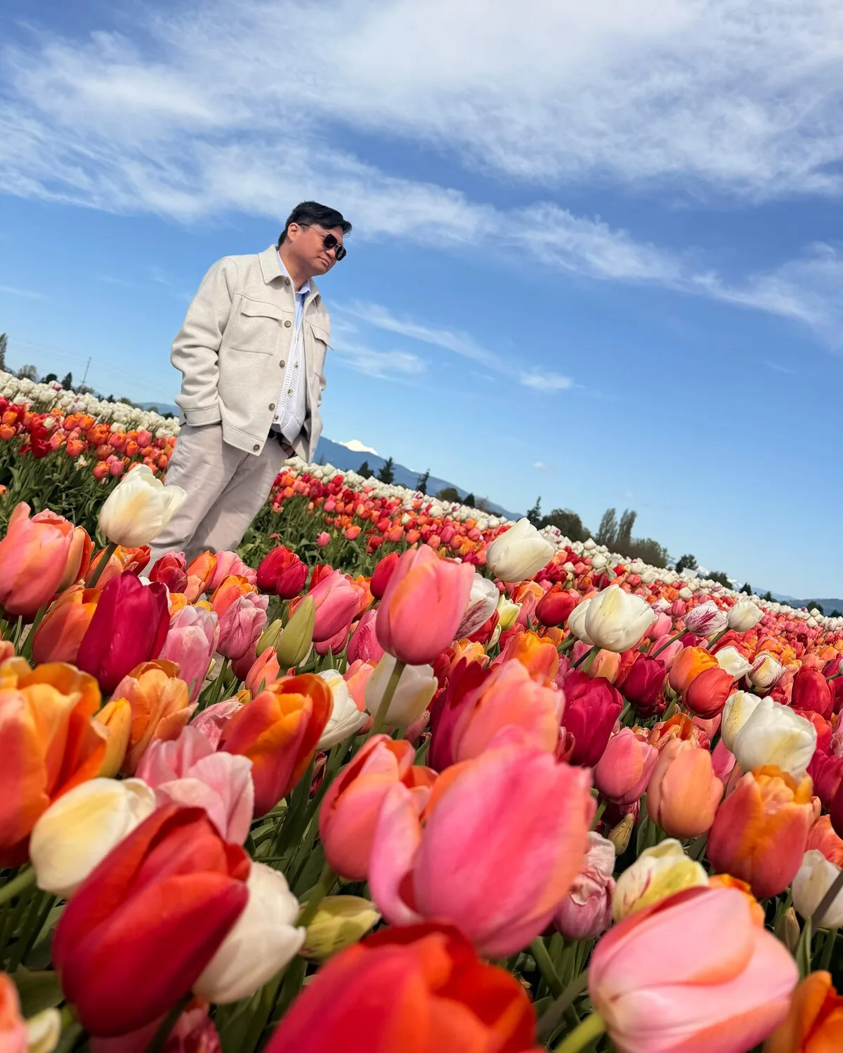 Warren de Leon in sunglasses and a cream jacket, standing in a field of red, pink and white tulips under a blue sky